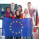 6 internationale Studierende stehen in einem Klassenzimmer, zeigen ihre Daumen hoch und halten eine Europa-Flagge