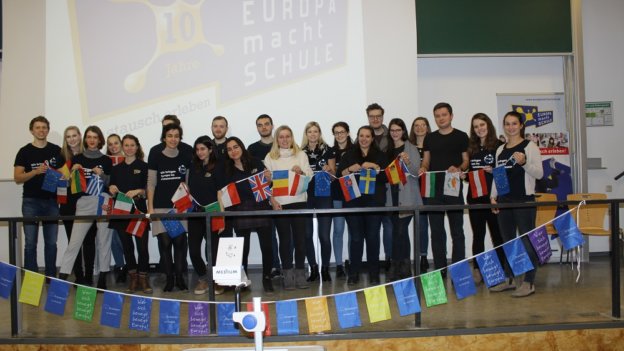 Europäische Gaststudierende und Standort-Team stehen gemeinsam auf der Bühne und halten eine Kette mit europäischen Flaggen.