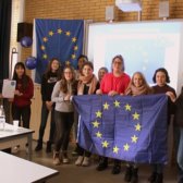Einige Schülerinnen und Schüler der Klasse sowie die Lehrerin und ein Standortmitglied stehen vorne in einem Klassenzimmer, halten eine Europaflagge und lachen in die Kamera.