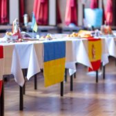 Aneinandergereihte Tische die mit einer weißen Tischdecke und einer europäischen Flagge geschmückt sind. Auf dem Tisch sind Ausstellungsstücke zu dem jeweiligen Land zu sehen.