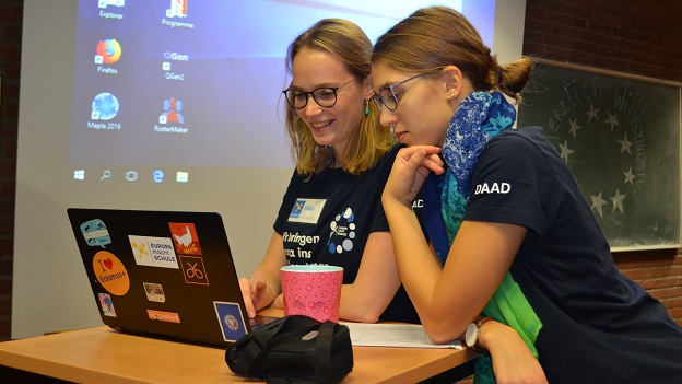 Zwei junge Frauen vom Standort-Team Hamburg sitzen, mit einem EmS-T-Shirt, vor einem Laptop und bereiten eine Präsentation für das EmS-Koordinationstreffenvor