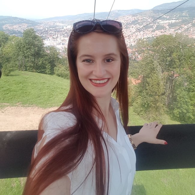 Frau Imamovic steht im Vordergrund. Im Hintergrund ist Sarajevo umgeben von einer grünen Landschaft zu sehen sowie die Trebevic Seilbahn.