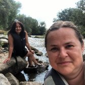Zwei EmS-Lehrkräfte sitzen zusammen an einem Fluss und schauen in die Kamera 