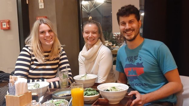 Drei junge Menschen lächeln und sitzen gemeinsam an einem Tisch mit Essen
