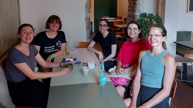 Fünf junge Frauen sitzen in einem Cafe an einem Tisch und lächeln in die Kamera