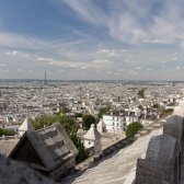 Paris fotografiert von einem Gebäude aus, dessen Dach man sieht. Der Himmel ist blau mit Wolkenstreifen und der Eiffelturm prägnant als Wahrzeichen zu erkennen.
