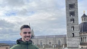 Ein Mann in einer grünen Jacke steht lächelnd an einem Geländer, im Hintergrund ist eine Kirche mit einem hohen Turm und schwarz-weißem Gestein zu sehen